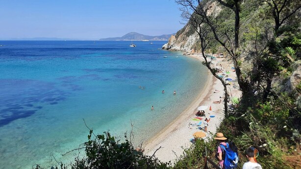 A coastal beach with clear turquoise waters, a sandy shore, and rocky cliffs.
