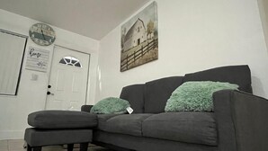 A living room with a gray sofa, two matching pillows, a small ottoman, a wall clock, a framed picture, and a door with a sign. - Gorgeous 3Bed in the heart of South Florida (Fort Lauderdale)