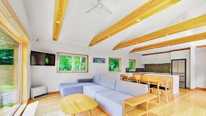 Un salon moderne avec un canapé d’angle, des poutres en bois, un coin repas avec une table et des chaises, et une cuisine en arrière-plan.
