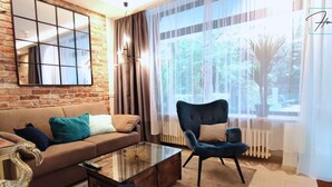 A living room with a brick wall, a sofa with cushions, a glass coffee table, a blue armchair, and a view of greenery through a window. - Homely Stay -  Studio P1 (München)