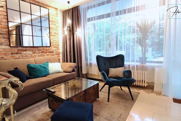 Un salon avec un mur en briques, un canapé avec des coussins, une table basse en verre, un fauteuil bleu et une vue sur la verdure par une fenêtre.
