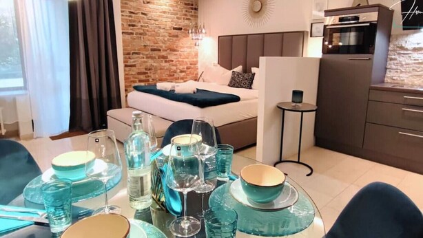 A modern hotel room with a brick wall, a bed with a headboard, a kitchenette, and a dining area set with blue glassware and plates. - Homely Stay -  Studio P1 (München)