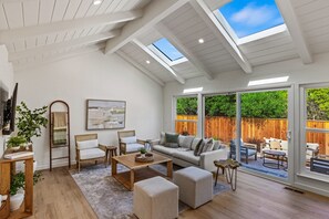 Ein geräumiges Wohnzimmer mit hohen Decken, Oberlichtern, einer Sofagarnitur, einem Couchtisch und Blick auf einen Holzzaun und Grünflächen draußen.