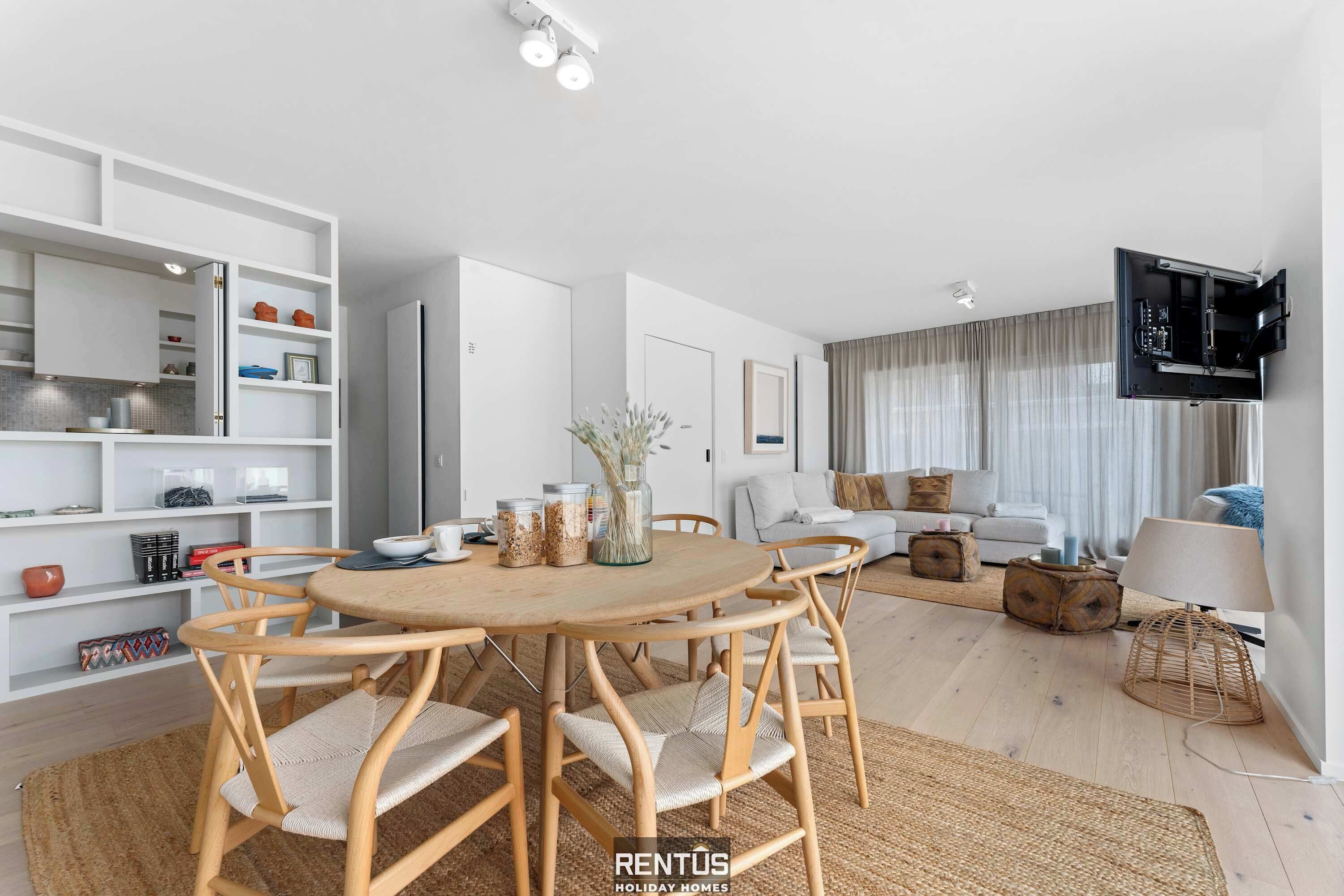 A modern living room with a round wooden dining table, white shelving, and a flat-screen TV mounted on the wall.