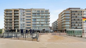 A multi-story residential building with balconies, a sandy ground area, and a glass-walled structure in the foreground. - Seaflow-holiday apartment with frontal sea view (Oostduinkerke)
