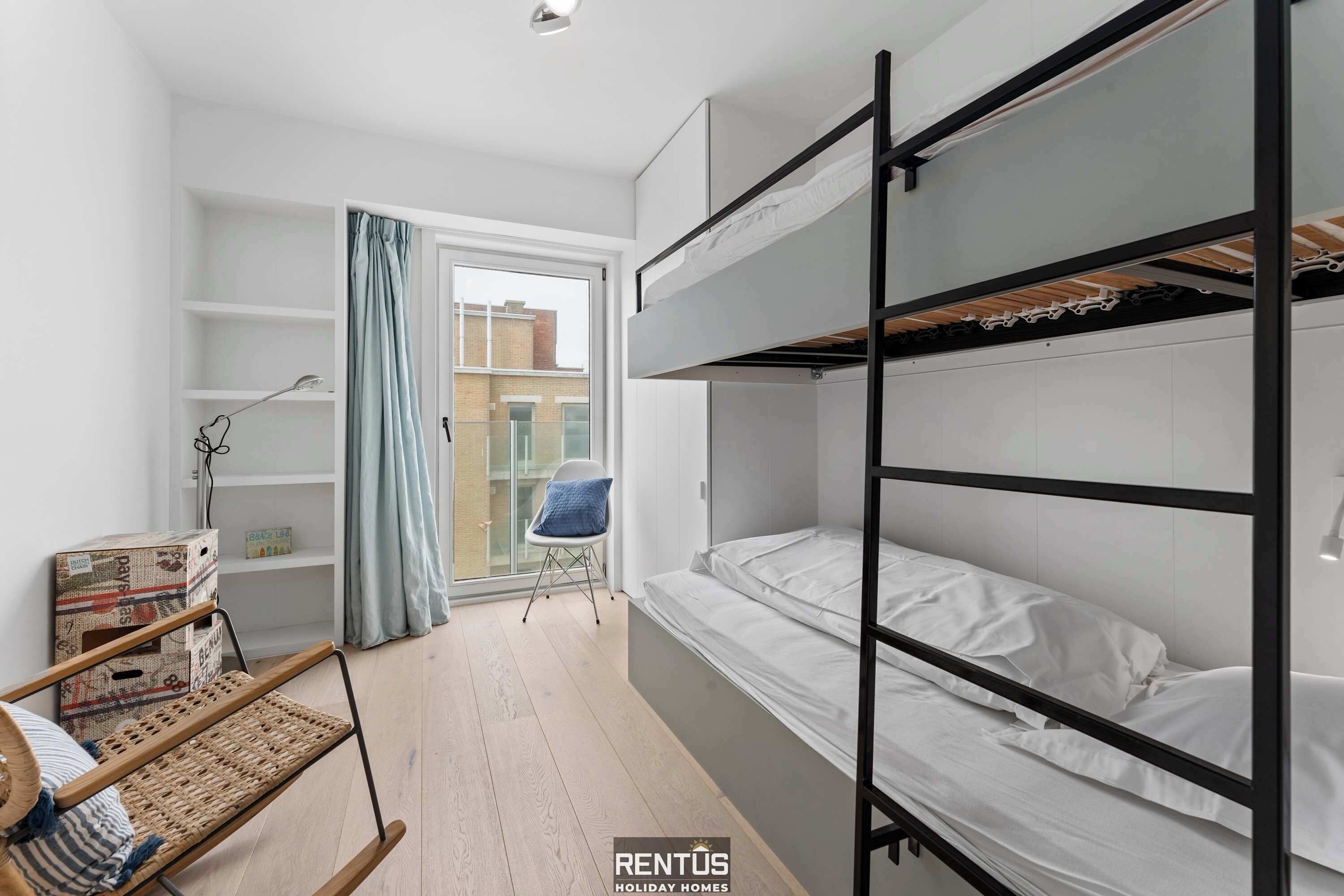 A compact bedroom with bunk beds, a small seating area, and a window with curtains.