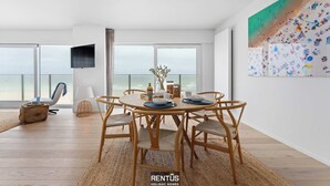 A spacious living area with a round wooden table, comfortable chairs, and a large window overlooking the beach. - Seaflow-holiday apartment with frontal sea view (Oostduinkerke)