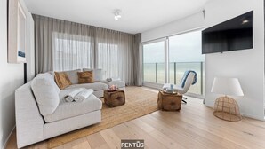 A modern living room with a sofa, two side tables, a lamp, and a large window with curtains. - Seaflow-holiday apartment with frontal sea view (Oostduinkerke)