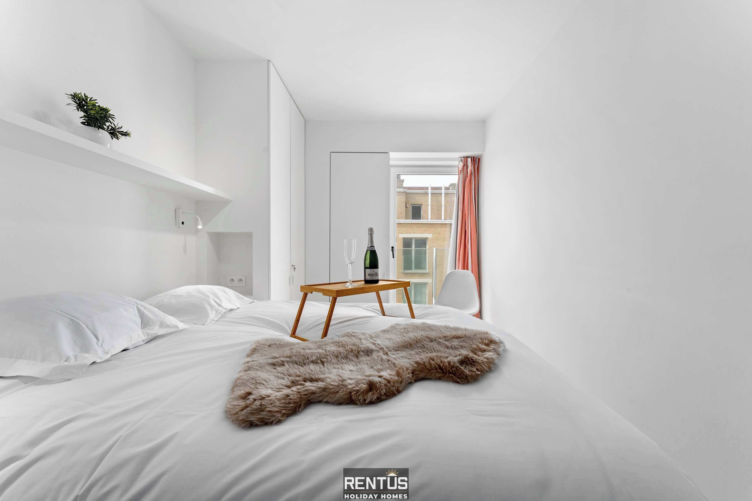 Et moderne hotelværelse med en seng, hvide vægge, et trænatbord, en champagneflaske og udsigt til bygninger.