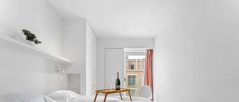 Et moderne hotelværelse med en seng, hvide vægge, et trænatbord, en champagneflaske og udsigt til bygninger.