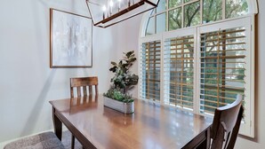 Uma área de refeições com uma mesa de madeira, cadeiras, um vaso de planta, um quadro na parede e uma janela com persianas.