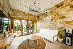 Relax in this stylish living room with a sofa against a natural stone wall, a TV, and a large window offering a view of lush greenery.