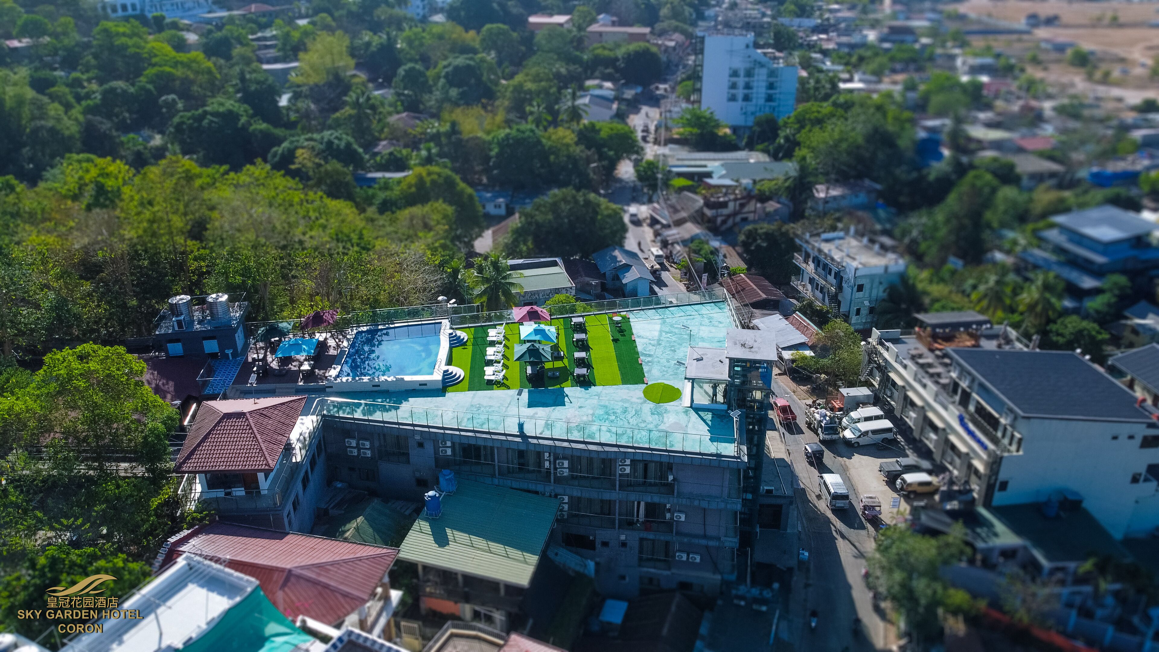 An aerial view of a hotel with a rooftop pool, surrounded by dense greenery and urban buildings.