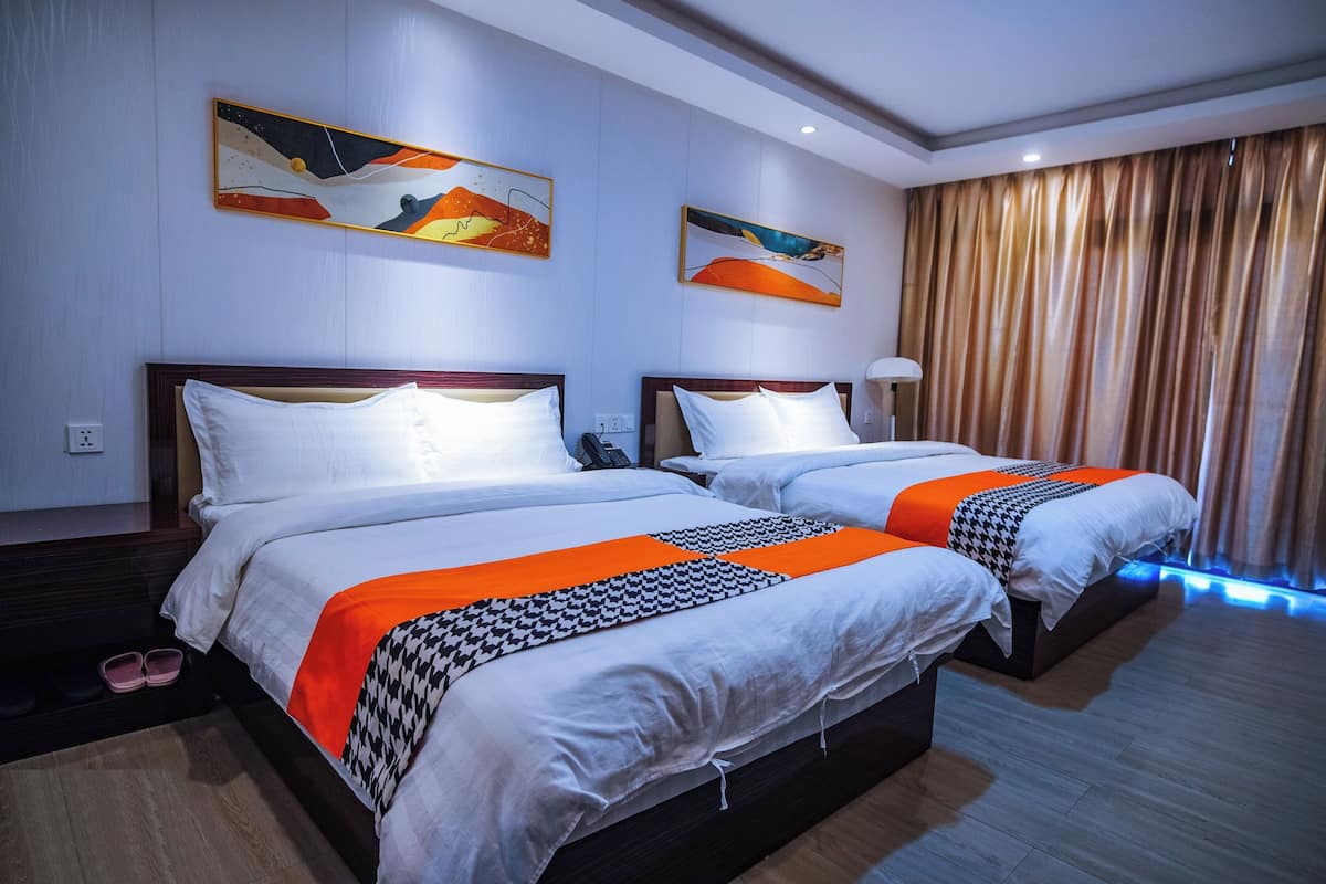 Quarto de hotel com duas camas, cada uma com roupa de cama branca e detalhes em laranja, uma luminária no teto e uma janela grande com cortinas.