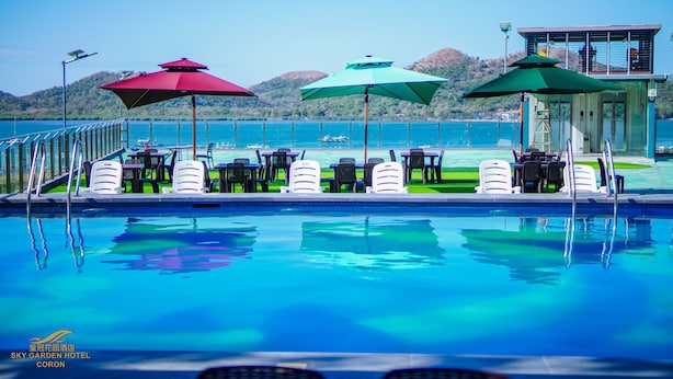 A poolside area with lounge chairs and umbrellas, a lifeguard tower, and a view of the sea and hills. - Sky Garden Hotel (Coron)