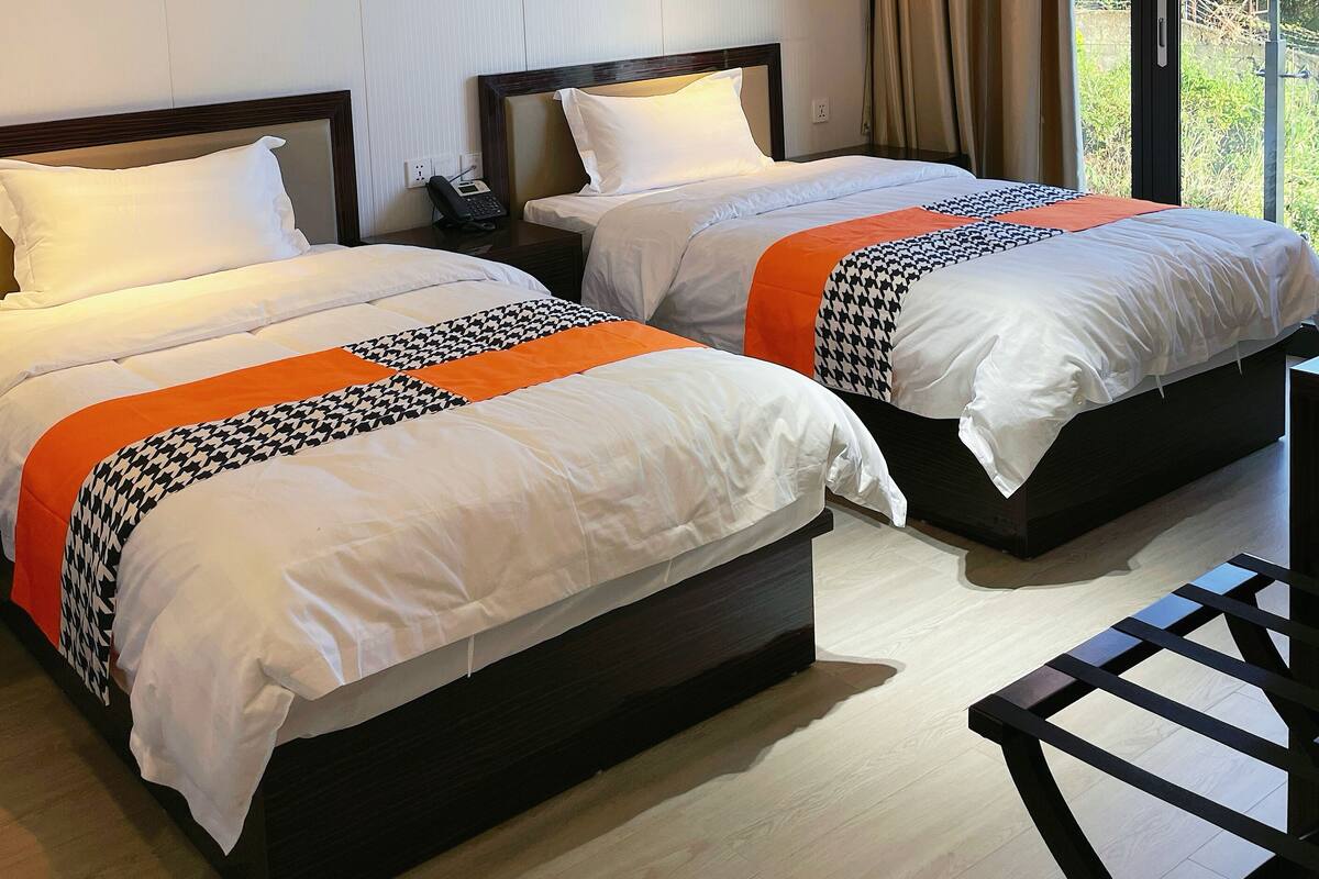 Duas camas de casal com roupa de cama branca e laranja, um telefone ao lado da cama e piso de madeira.