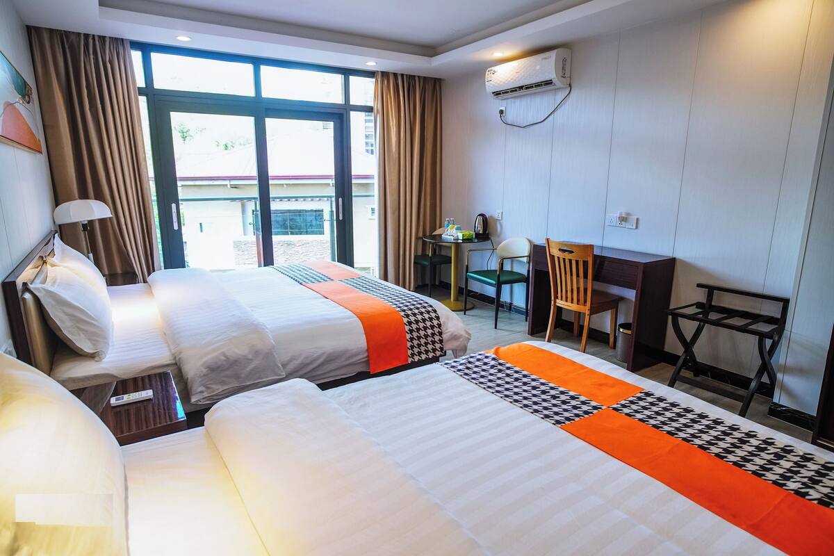 Quarto de hotel com duas camas, uma escrivaninha, uma cadeira e vista para o exterior.