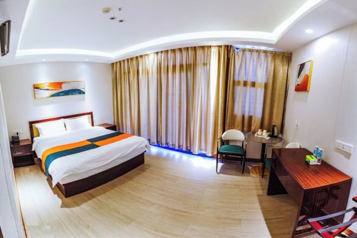 A hotel room with a bed, a desk, a chair, and a view of the outside through curtains.