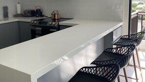 A modern kitchen with white cabinetry, a stainless steel range hood, a microwave, and a gas stove. There are four black bar stools with wooden legs.