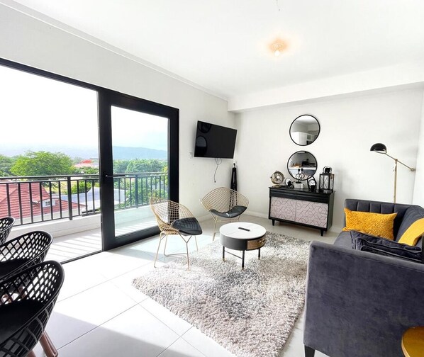 Ein modernes Wohnzimmer mit Balkon, einem Flachbildfernseher, einem runden Spiegel und einem Sofa mit gelbem Kissen.