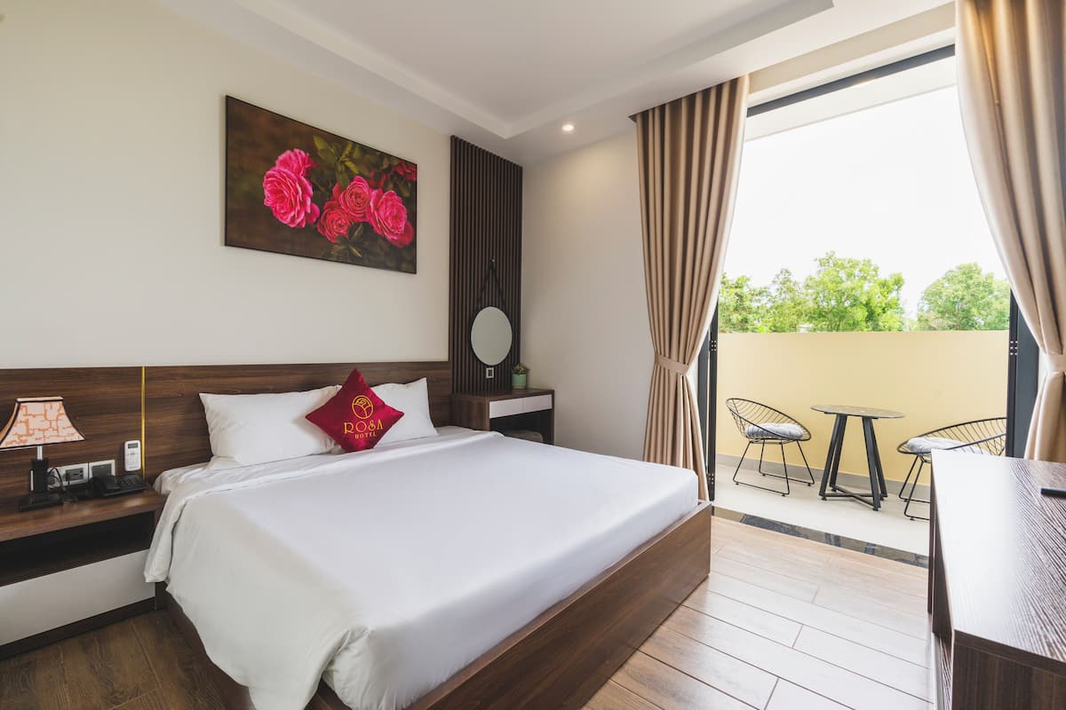 A hotel room with a large bed, a bedside table with a lamp, a painting of roses, a round mirror, and a view of the outside balcony with chairs.