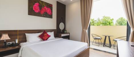 Habitación de hotel con una cama grande, una mesita de noche con lámpara, un cuadro de rosas, un espejo redondo y vistas al balcón exterior con sillas.