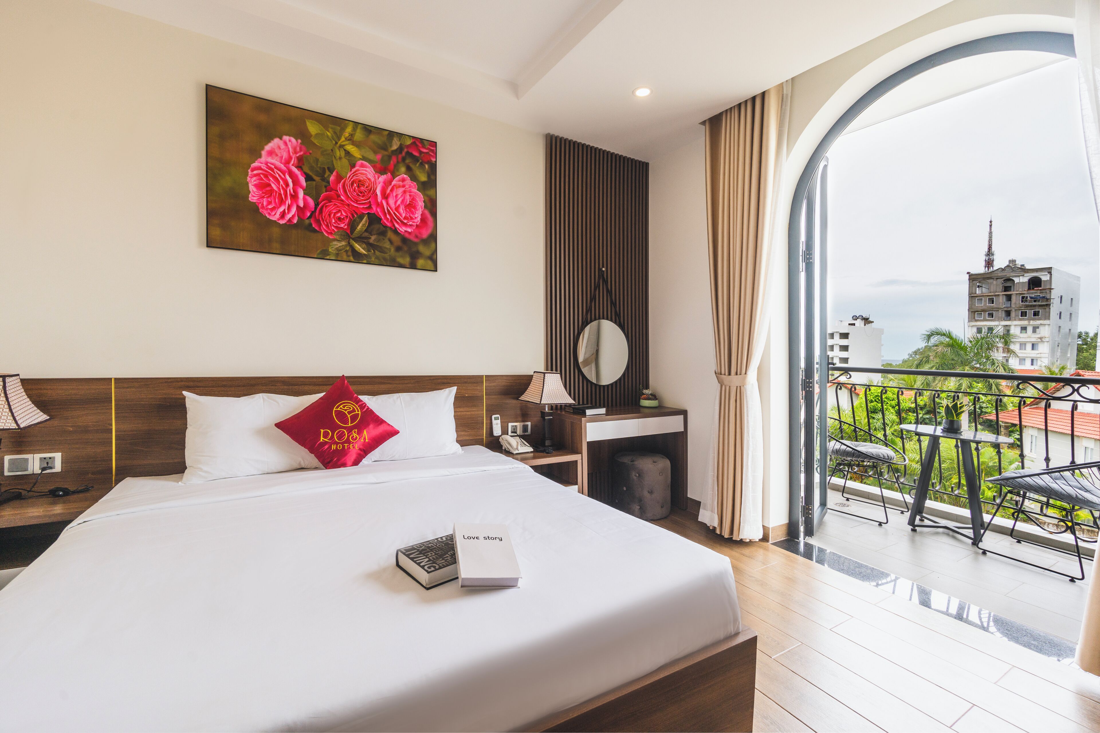 A hotel room with a large bed, a bedside table with a lamp, a mirror, and a view of the city through an open balcony.
