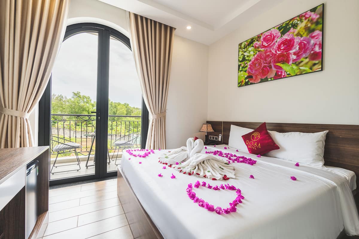 A hotel room with a balcony, a bed with white linens, a red pillow, and a painting of pink flowers on the wall.