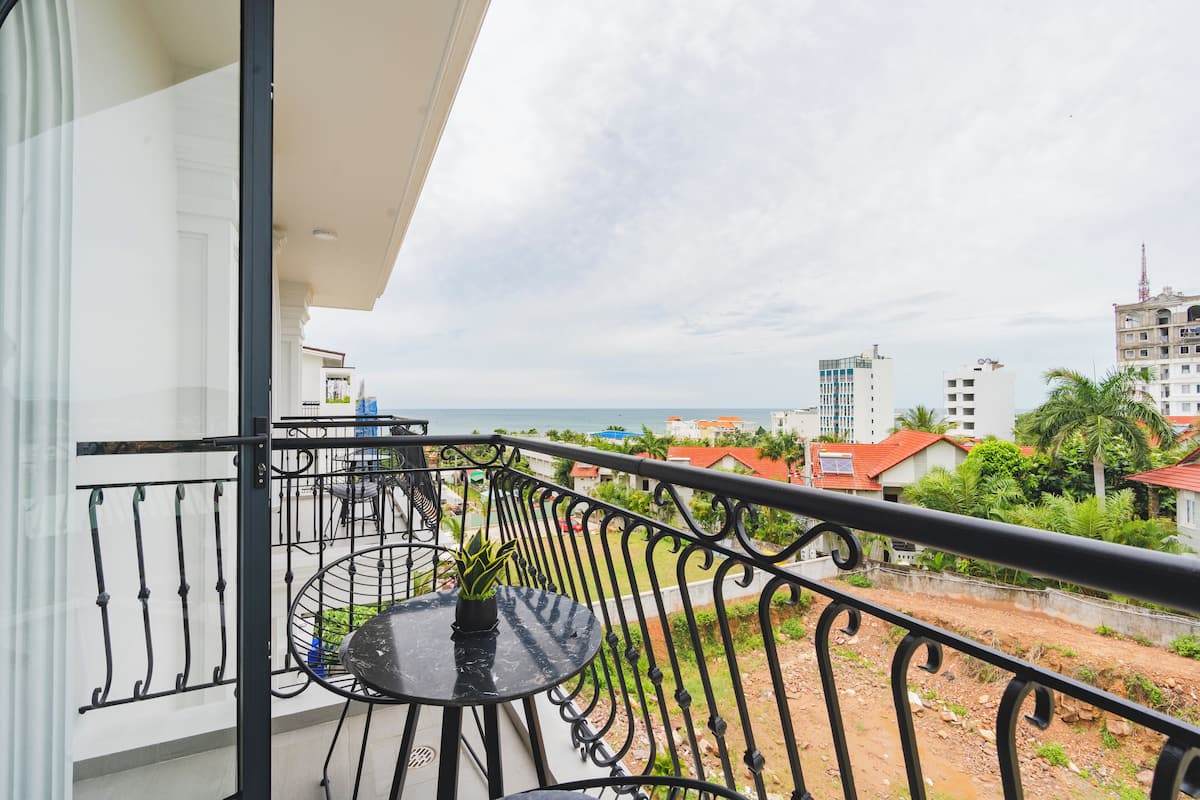A balcony with a round table and potted plant, overlooking a coastal area with buildings and greenery.