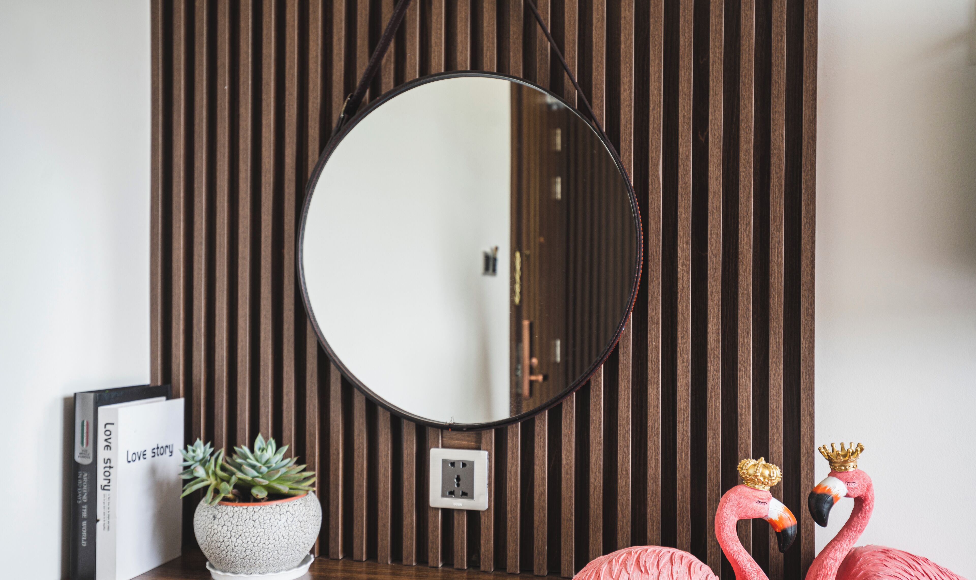 A round mirror on a wall with vertical wooden slats, a potted succulent, a book titled 'Love story', and two flamingo figurines with crowns.