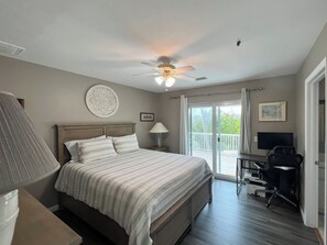 A bedroom with a bed, a desk, a chair, a ceiling fan, and a balcony with a view.