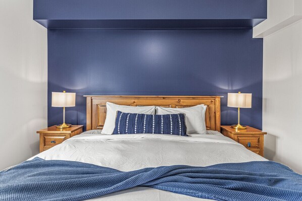 Een slaapkamer met een houten hoofdbord, twee nachtkastjes met lampen, een bed met wit beddengoed en een blauwe accentmuur.