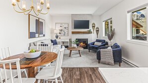 A modern living room with a fireplace, a dining area, and a large window offering a mountain view. - Top Floor Mountain View with Pool & Hot tubs (Canmore)
