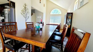 Een eetruimte met een houten tafel en stoelen, een keuken op de achtergrond, en een groot raam met uitzicht op bomen.