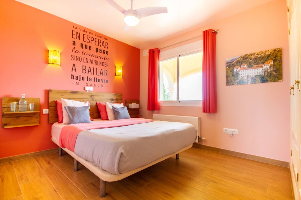 Une chambre à coucher comprenant un lit, une tête de lit en bois, des tables de chevet, une étagère fixée au mur, une fenêtre avec des rideaux et un mur orné d’une citation inspirante et d’un tableau.