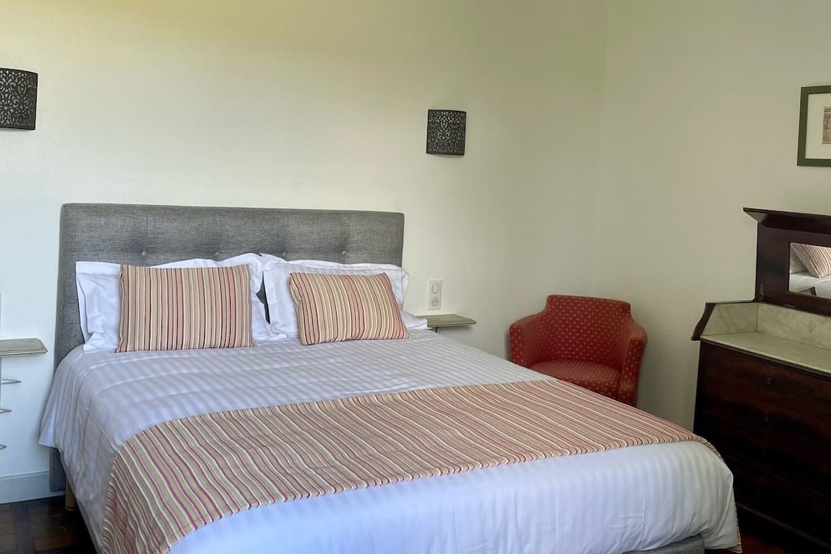 Un lit bien fait, avec une literie à rayures, une chaise rouge et une commode en bois, dans une pièce simple aux murs unis.