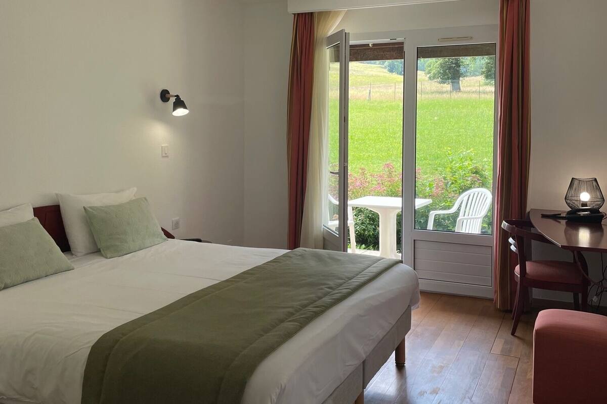 Une chambre à coucher avec un grand lit, un bureau avec une lampe et une vue sur un jardin, visible par une porte ouverte.