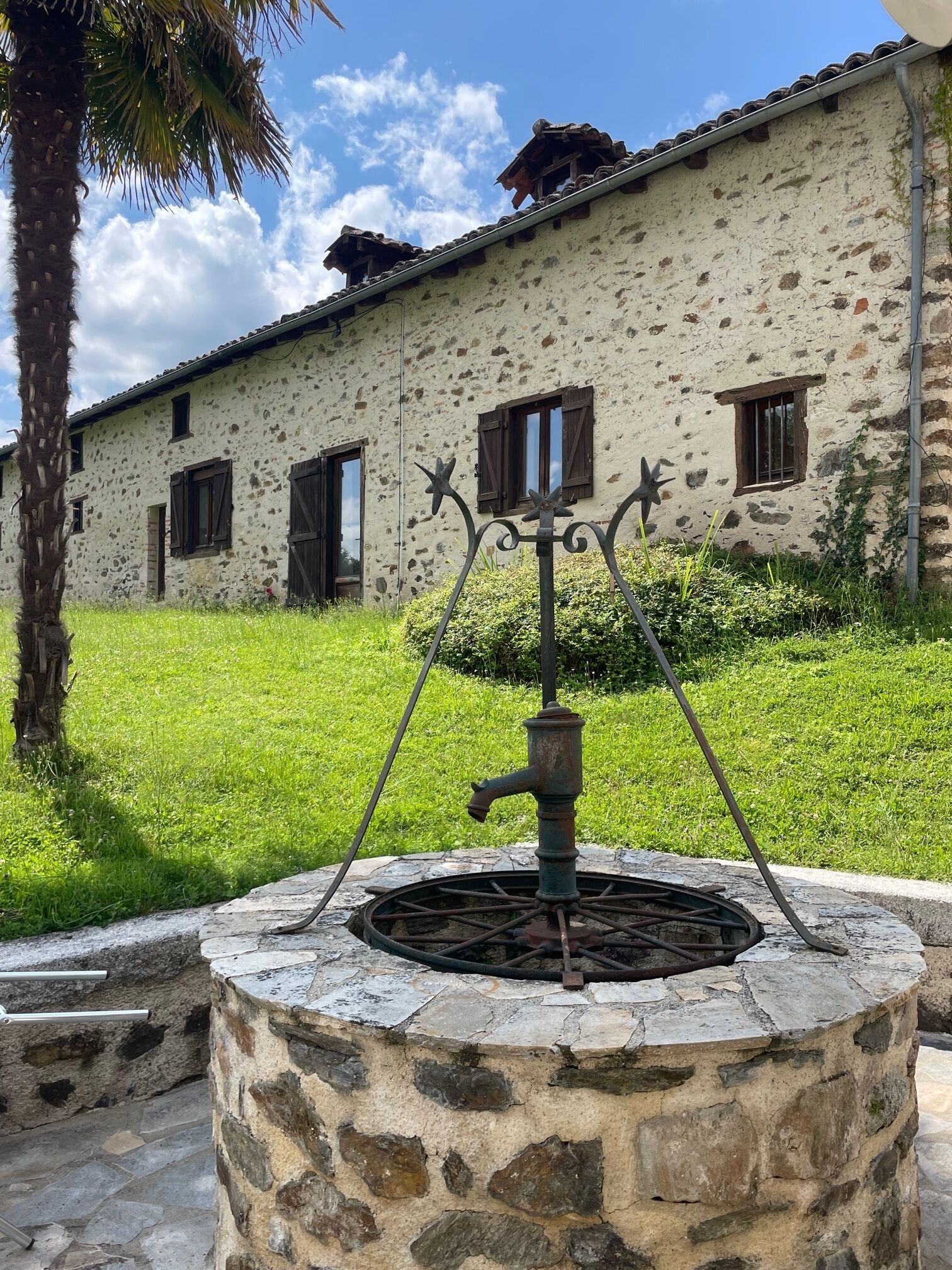 A rustic stone well with a metal spout, surrounded by a stone wall, in front of a building with a stone facade and wooden shutters.