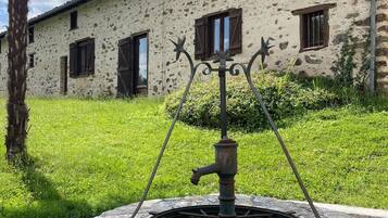 A rustic stone well with a metal spout, surrounded by a stone wall, in front of a building with a stone facade and wooden shutters.