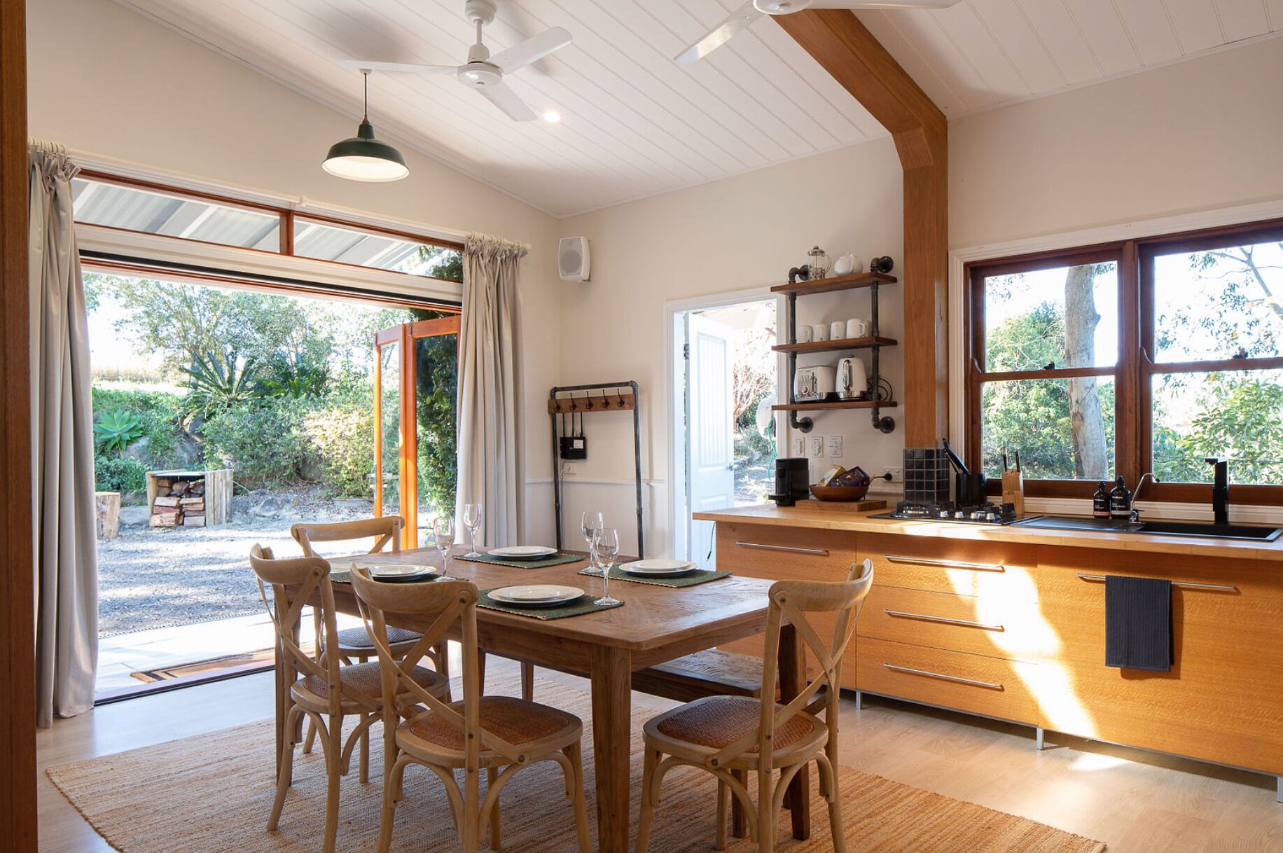 A dining area with a wooden table and chairs, a kitchen with a sink and shelves, and a view of the outdoors through large windows.