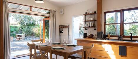 A dining area with a wooden table and chairs, a kitchen with a sink and shelves, and a view of the outdoors through large windows.