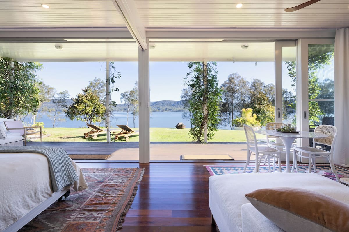 A modern bedroom with a large window overlooking a lake and hills. There is a bed, a small table, and a chair.