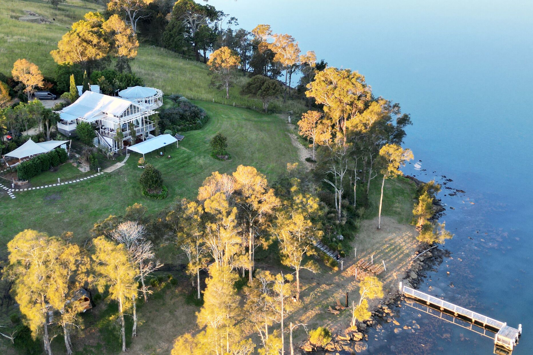 An aerial view of a coastal property with a large house, a dock, and a bridge leading to a pier.