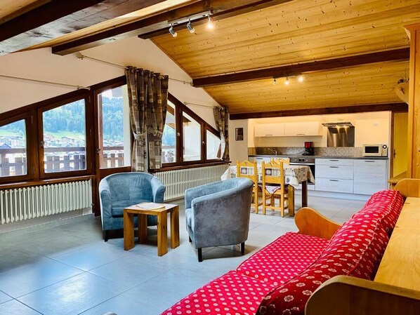 Um quarto acolhedor com teto de madeira, um sofá, duas poltronas, uma pequena mesa e vista para uma cozinha e área externa através de amplas janelas.