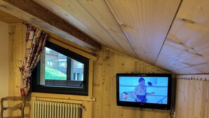 Une pièce aux murs lambrés, avec un téléviseur fixé au mur, une fenêtre donnant sur des immeubles, un radiateur et une chaise en bois.