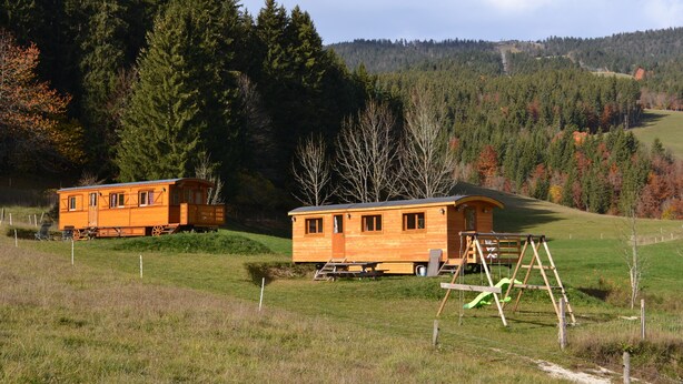 Two wooden cabins in a grassy field with a playground and a forested hill in the background. - Roulottes a la Ferme sur le Plateau du Vercors (Autrans-Méaudre en Vercors)
