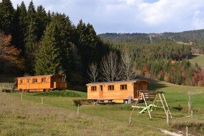 Two wooden cabins in a grassy field with a playground and a forested hill in the background. - Roulottes a la Ferme sur le Plateau du Vercors (Autrans-Méaudre en Vercors)