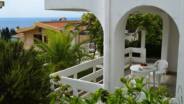 A balcony with white railings, a round table with chairs, and a view of the sea and buildings in the background. - Apartment for 4 persons (Bar)
