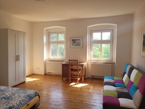 Ein Zimmer mit einem Bett, einem Holztisch, zwei Fenstern, einem Heizkörper und einem farbenfrohen Sofa.