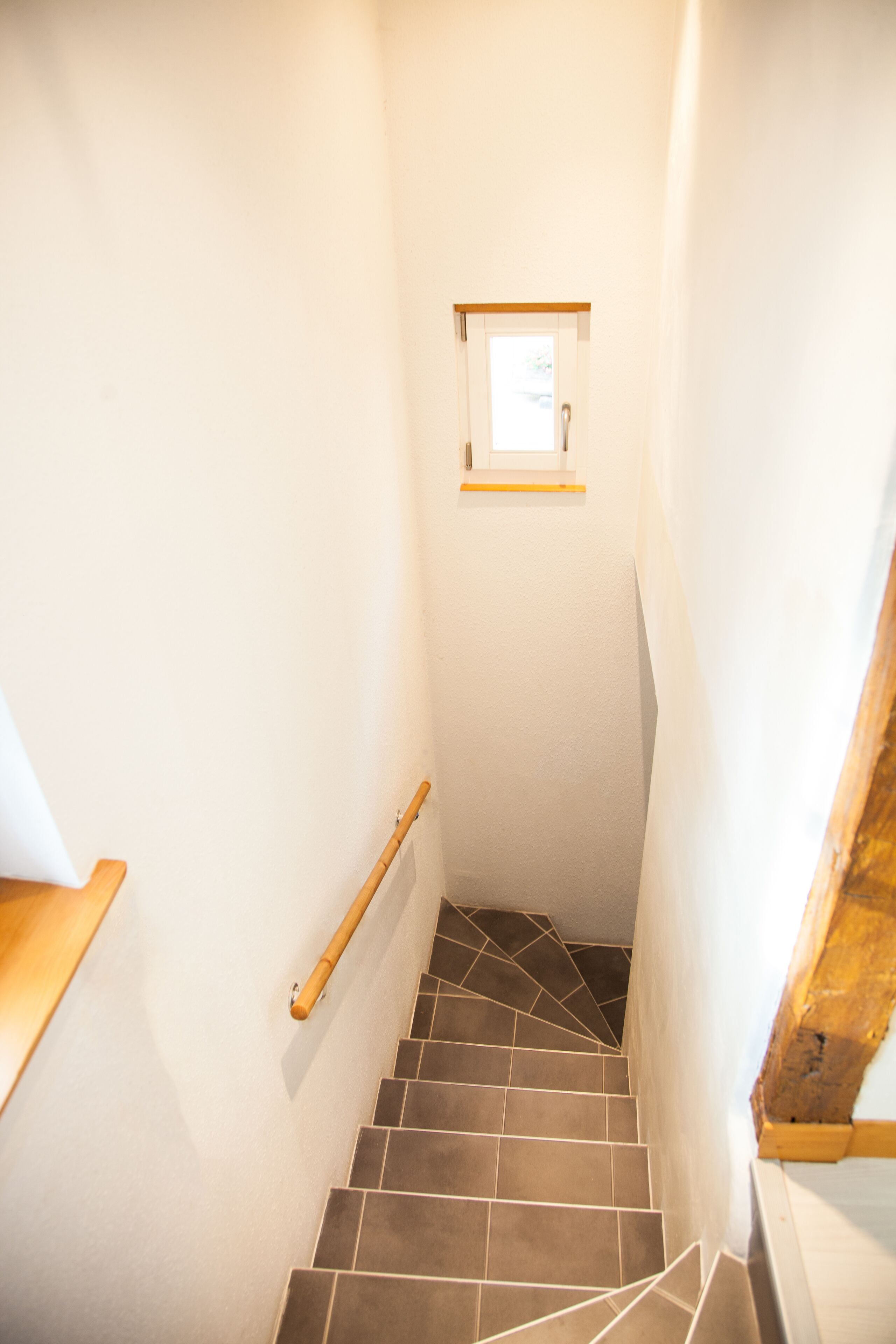 Un escalier étroit doté d’une main courante, une petite fenêtre en haut et un cadre de porte en bois.
